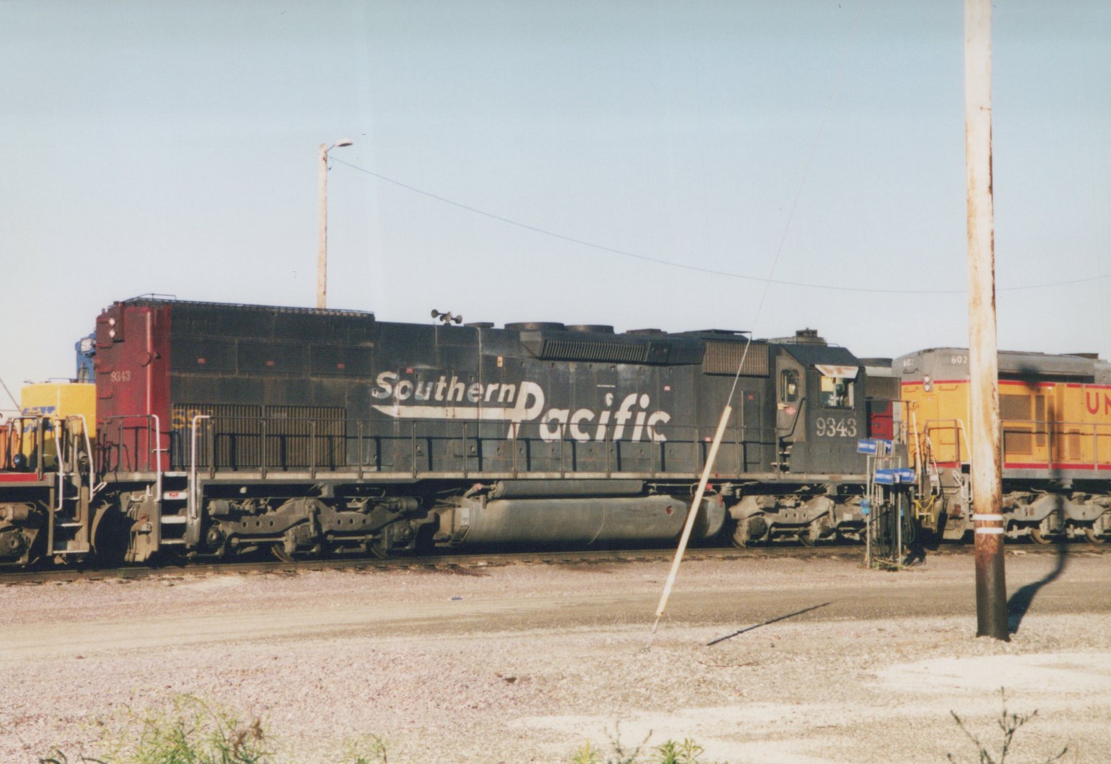 A black Southern Pacific locomotive with the number 9343 is parked next to a yellow Union Pacific locomotive with the number 602 in a gravel yard under a clear sky.