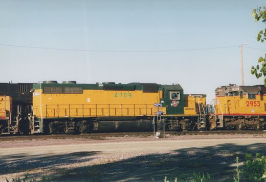 A side view of two yellow and green diesel locomotives, numbered 4709 and 2953, parked on parallel tracks in a gravel yard with trees and a clear blue sky in the background.