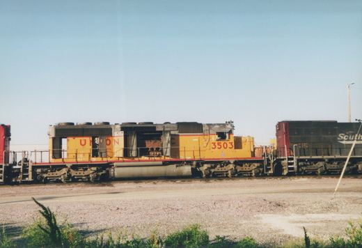 A yellow and red Union Pacific locomotive, number 3503, is seen from the side in a gravel rail yard on a clear day, with another dark locomotive coupled behind it.