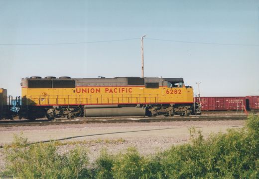 Side view of a yellow Union Pacific locomotive numbered 6282 pulling red Wisconsin Central boxcars in a gravel rail yard under a clear sky.