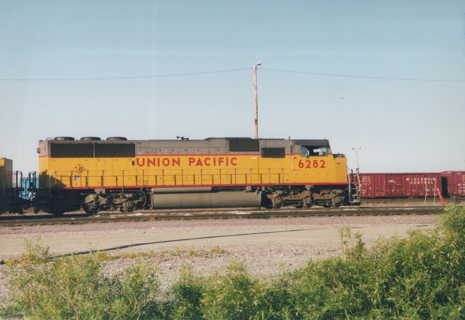 Side view of a yellow Union Pacific locomotive numbered 6282 pulling red Wisconsin Central boxcars in a gravel rail yard under a clear sky.