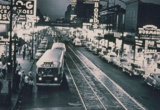 A CTA bus travels down a bustling Chicago street lined with vintage cars and illuminated signs, with people boarding and walking on the sidewalk.