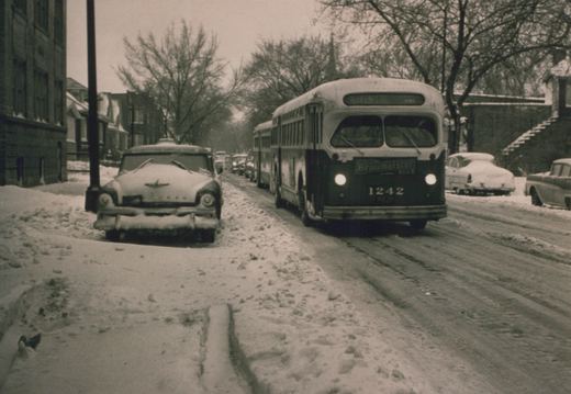 A black and white photo of a Chicago Transit Authority bus number 1242 driving down a snow-covered street lined with parked cars and bare trees.