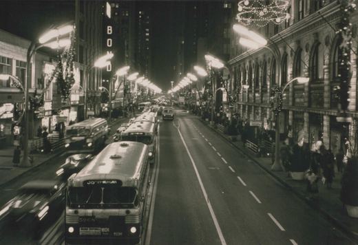 A black-and-white night scene of a city street with several buses, including a lead bus numbered 5968, surrounded by holiday lights and storefronts with people on the sidewalks.