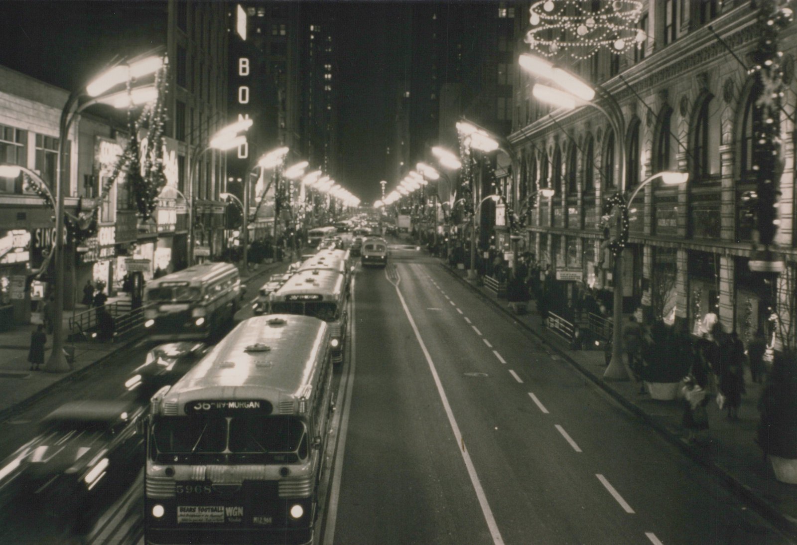 A black-and-white night scene of a city street with several buses, including a lead bus numbered 5968, surrounded by holiday lights and storefronts with people on the sidewalks.