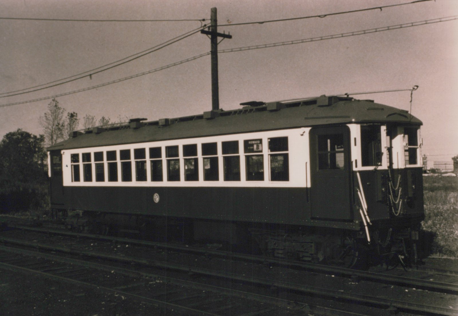 A black and white photograph of a vintage interurban car, numbered 4, with a destination sign reading 'HOWARD B', on a set of tracks with overhead wires and a wooden utility pole in the background.