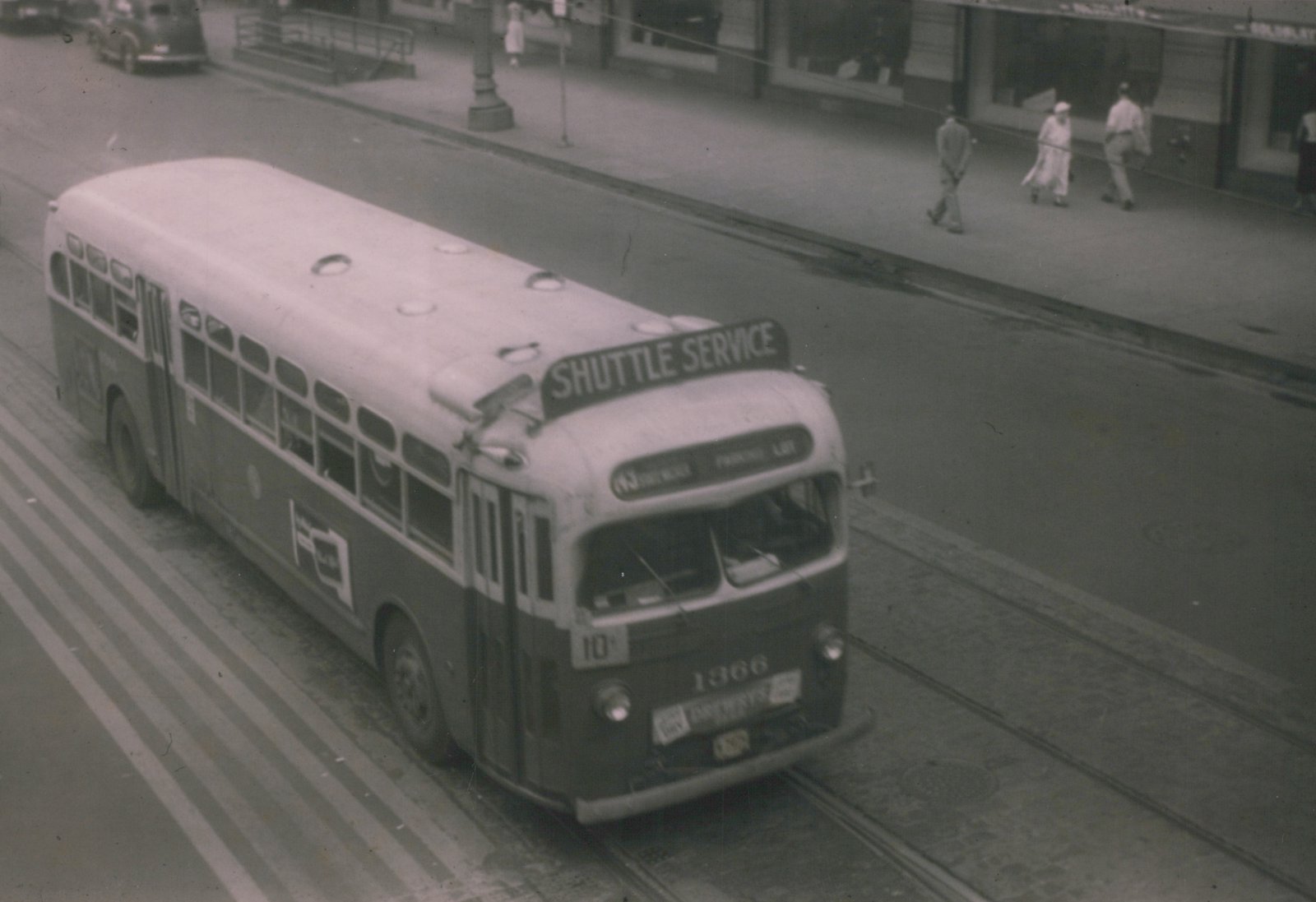 A vintage black-and-white photo of a two-tone GMC bus with 'SHUTTLE SERVICE' on its roof and the number 1366 on its front, driving on a street with trolley tracks and pedestrians on the sidewalk.