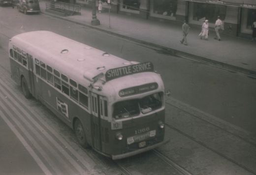 A vintage black-and-white photo of a two-tone GMC bus with 'SHUTTLE SERVICE' on its roof and the number 1366 on its front, driving on a street with trolley tracks and pedestrians on the sidewalk.