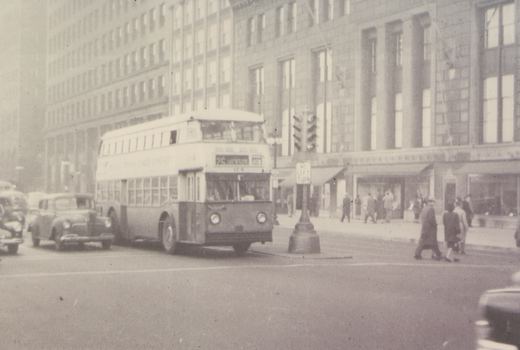 A black-and-white photo of a Chicago Motor Coach Company double-decker bus, number 114, on route 25 Henson, turning a corner in a city street with pedestrians and 1940s cars nearby.