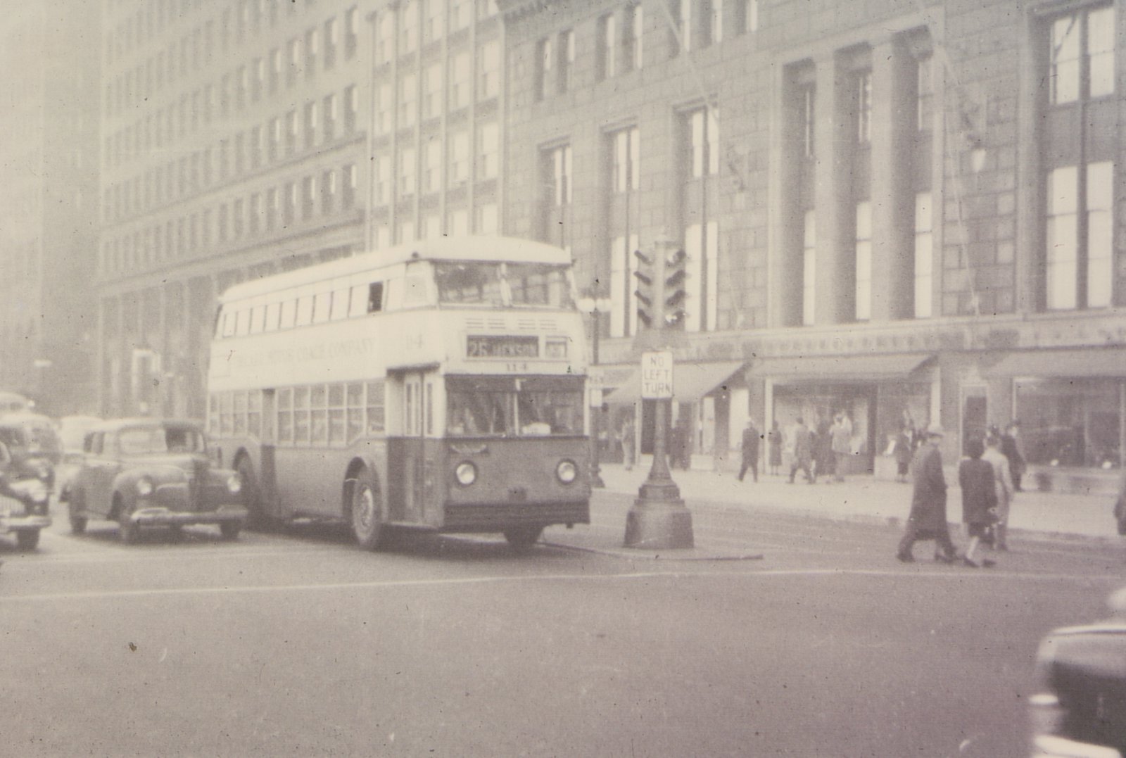A black-and-white photo of a Chicago Motor Coach Company double-decker bus, number 114, on route 25 Henson, turning a corner in a city street with pedestrians and 1940s cars nearby.