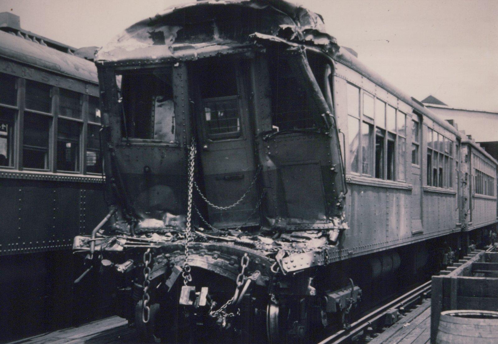 A black and white photo of a heavily damaged rapid transit car with its front end crumpled, sitting on an elevated track next to another car.