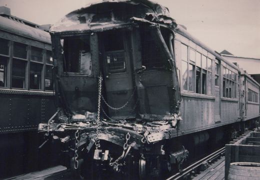 A black and white photo of a heavily damaged rapid transit car with its front end crumpled, sitting on an elevated track next to another car.