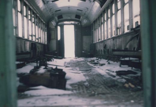 The interior of a abandoned rail car is filled with snow, broken seats, and debris, viewed from one end looking toward a bright doorway.