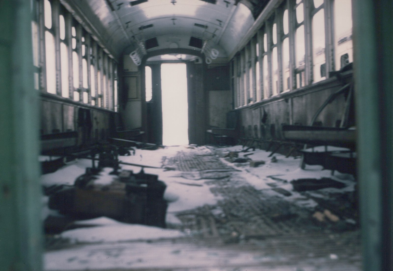 The interior of a abandoned rail car is filled with snow, broken seats, and debris, viewed from one end looking toward a bright doorway.