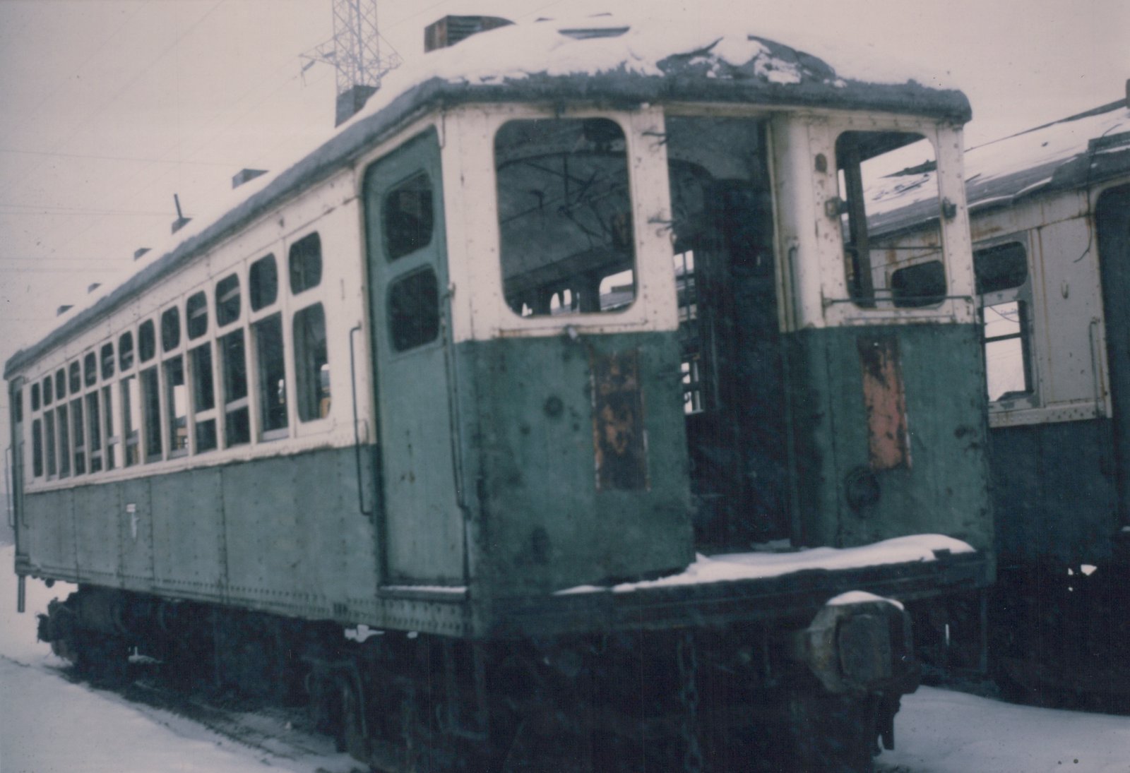 An old, dilapidated green and cream interurban car covered in snow, with broken windows and a rusted body, rests on tracks in a quiet, empty yard.