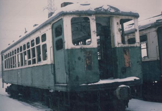 An old, dilapidated green and cream interurban car covered in snow, with broken windows and a rusted body, rests on tracks in a quiet, empty yard.