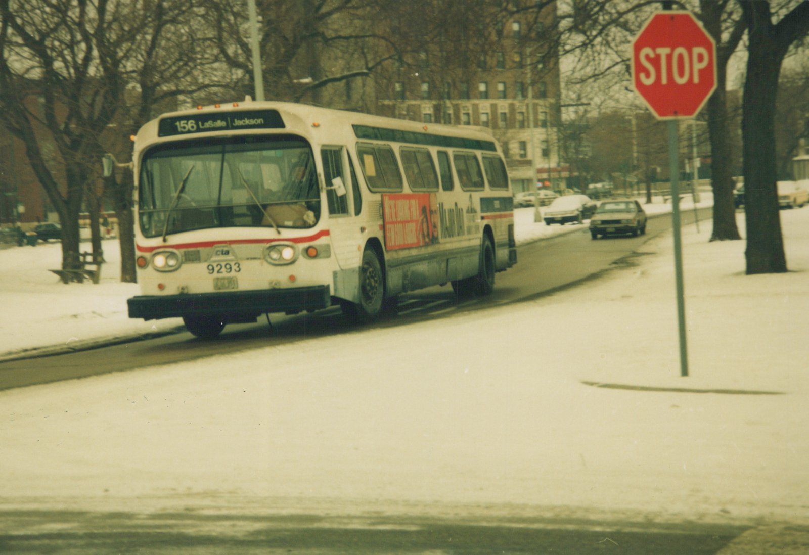 A white and red CTA bus with number 9293 is driving on a snow-covered street past a stop sign, with bare trees and buildings in the background.