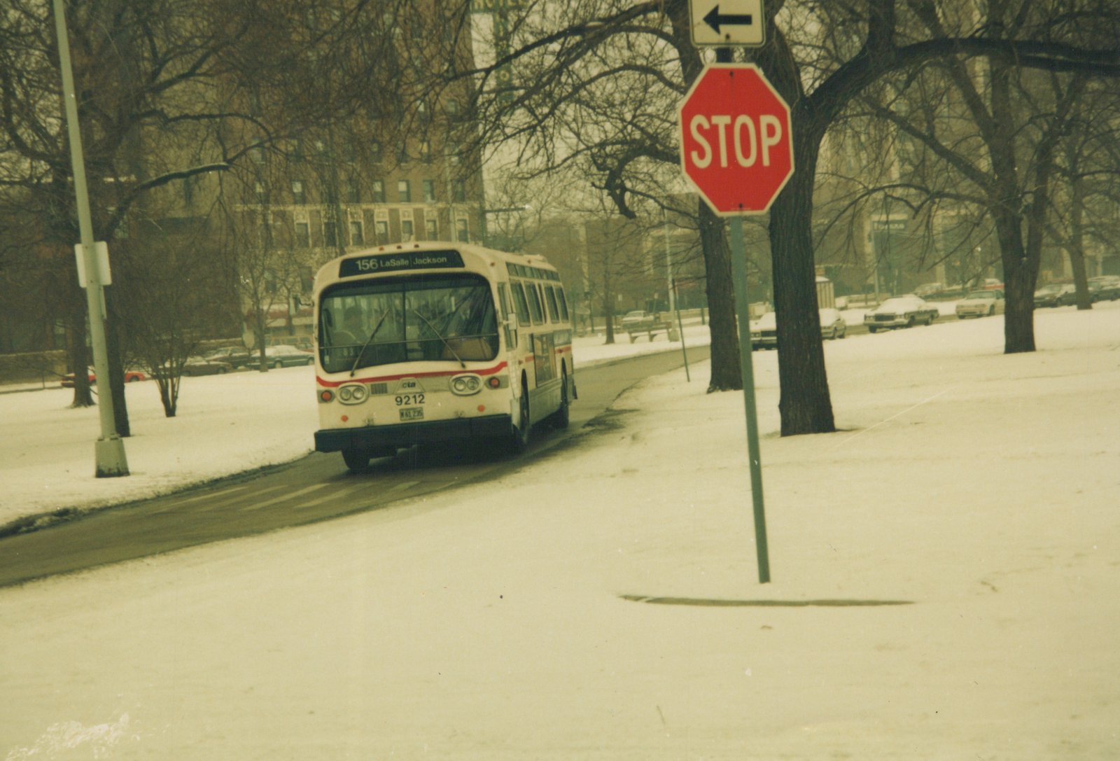 A white Chicago Transit Authority bus with red stripe, number 9212, drives on a snow-covered street past a stop sign, with snow-covered trees and buildings in the background.