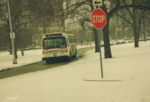 A white Chicago Transit Authority bus with red stripe, number 9212, drives on a snow-covered street past a stop sign, with snow-covered trees and buildings in the background.