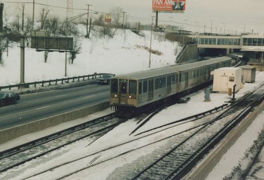 A tan and gray Chicago Transit Authority train with the number 3203 and destination 'O'Hare' is on an elevated snowy track, with a highway and a Pan Am billboard nearby.