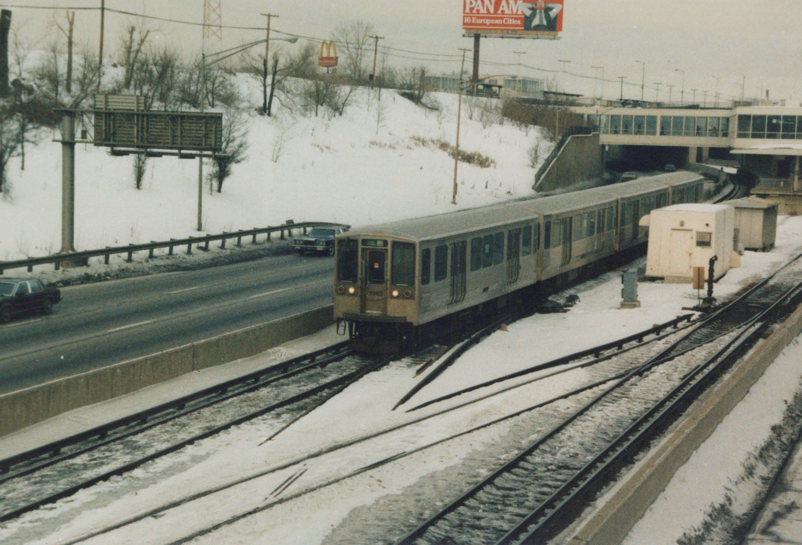 A tan and gray Chicago Transit Authority train with the number 3203 and destination 'O'Hare' is on an elevated snowy track, with a highway and a Pan Am billboard nearby.