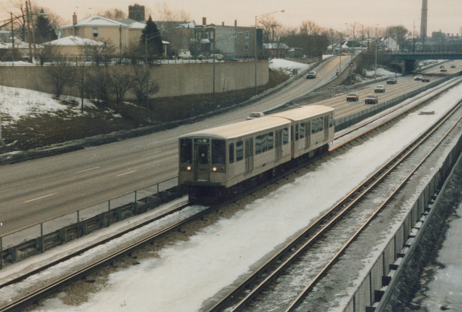 Two white Chicago 'L' cars travel on snow-dusted tracks next to a highway with cars driving on it; the lead car's number 2226 and route 22 are visible.