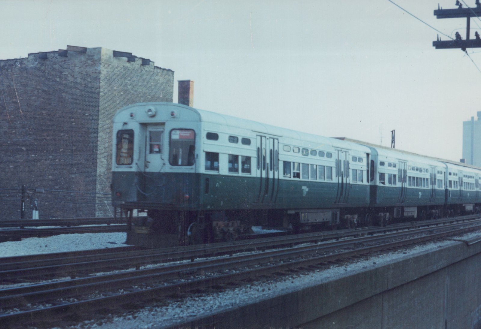 A side view of a green and cream South Shore Line train running on an elevated track next to a brick building under an overcast sky.