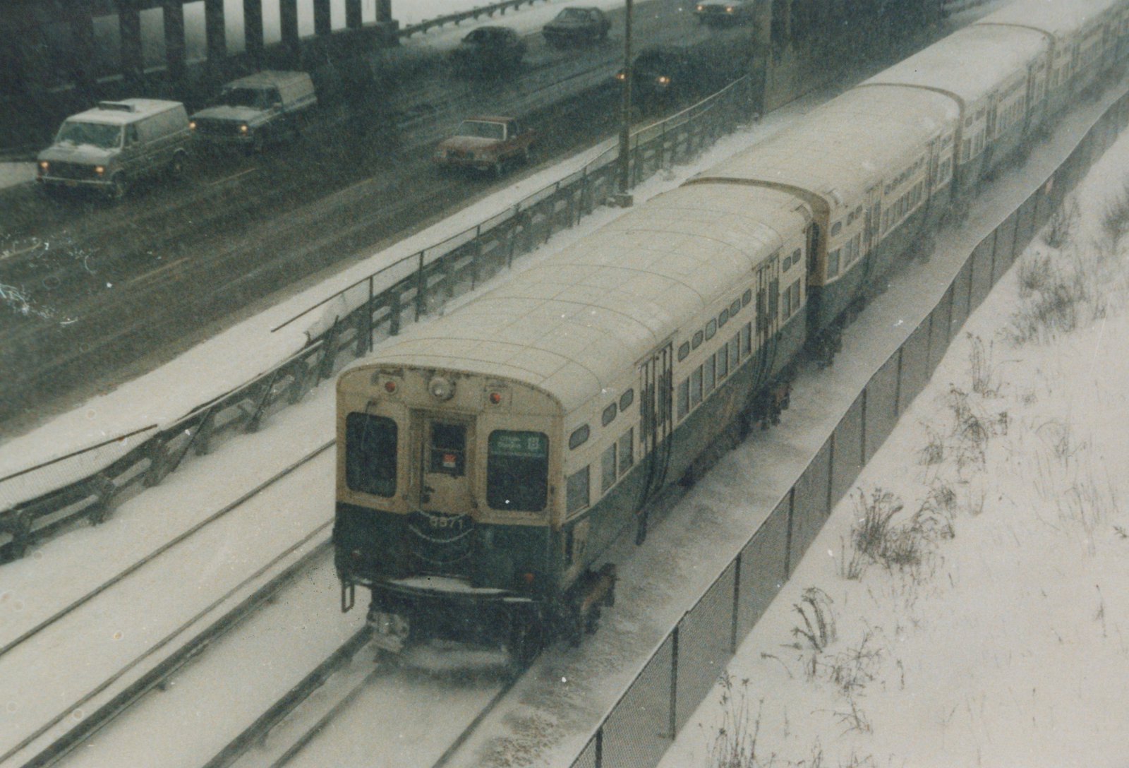 A cream and green Chicago Transit Authority train labeled 8971 is on elevated tracks next to a snowy highway with cars.