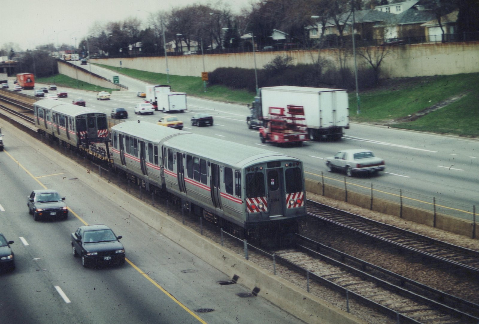 A silver Chicago Transit Authority train with red stripes travels on tracks in the median of a multi-lane highway, with cars and trucks visible on the road.