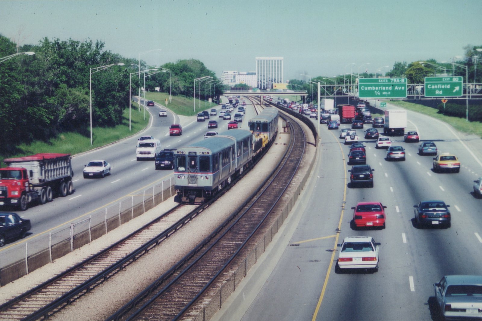 An elevated view of a Chicago Transit Authority train running on tracks in the median of a busy highway flanked by cars and trucks under a clear sky.