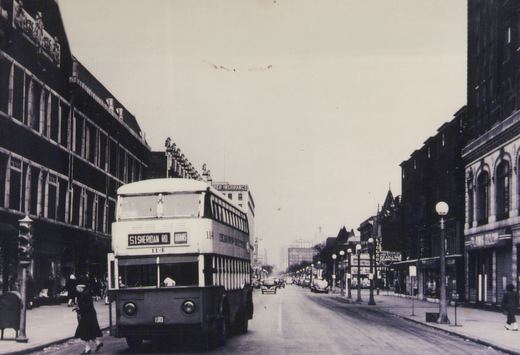 A black and white photo of a double-decker bus on a city street with pedestrians and old buildings. The bus is marked '51 SHERIDAN RD.' and 'ADAMS' with the number 114.