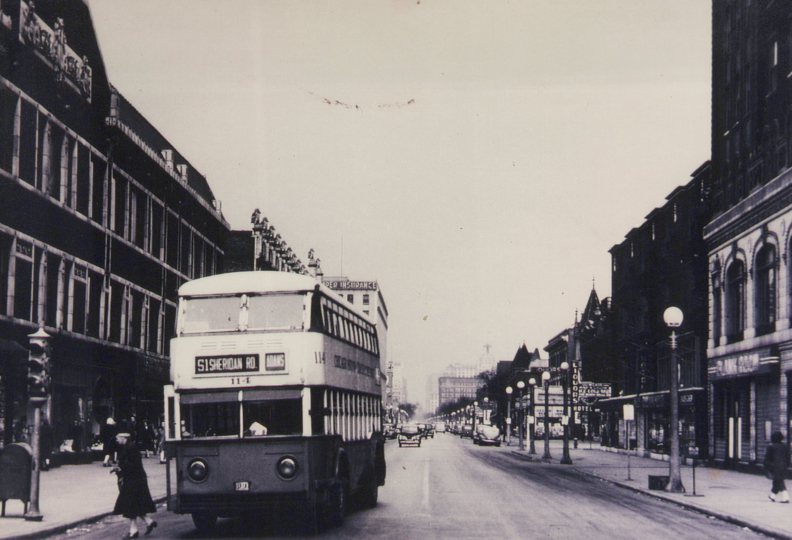 A black and white photo of a double-decker bus on a city street with pedestrians and old buildings. The bus is marked '51 SHERIDAN RD.' and 'ADAMS' with the number 114.