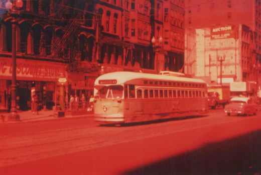 A red-tinted photograph of a vintage streetcar on a city street, with storefronts and other cars in the background.