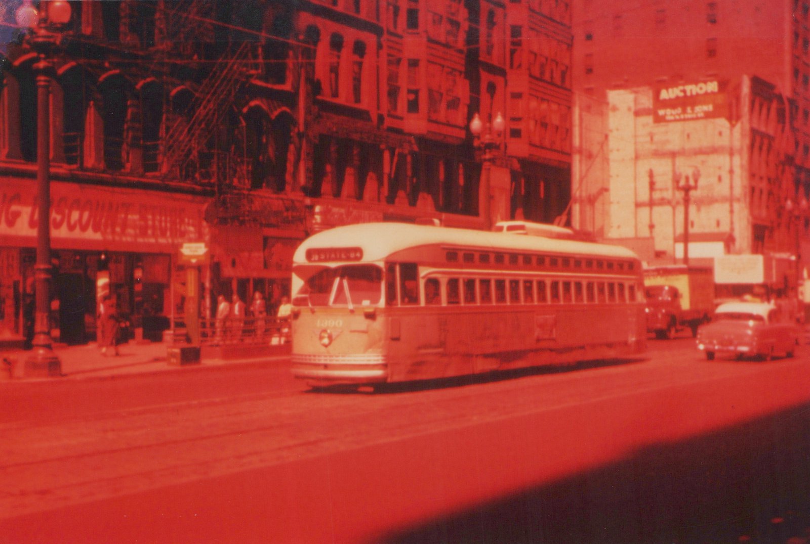 A red-tinted photograph of a vintage streetcar on a city street, with storefronts and other cars in the background.