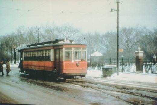 A red and cream Chicago Surface Lines streetcar, number 328, is stopped on a snowy street with bare trees in the background, as a few people wait to board.