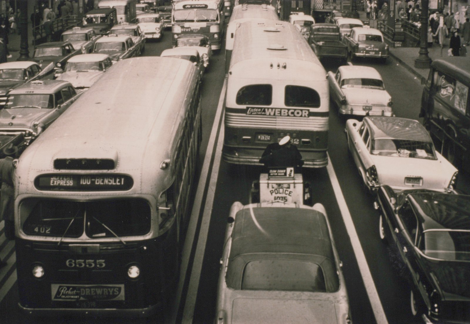 A high-angle black and white photograph of a crowded Chicago street, featuring a white Chicago Transit Authority bus numbered 6555 on the Jeffery Express route, another CTA bus, a police motorcycle, and numerous 1950s automobiles.
