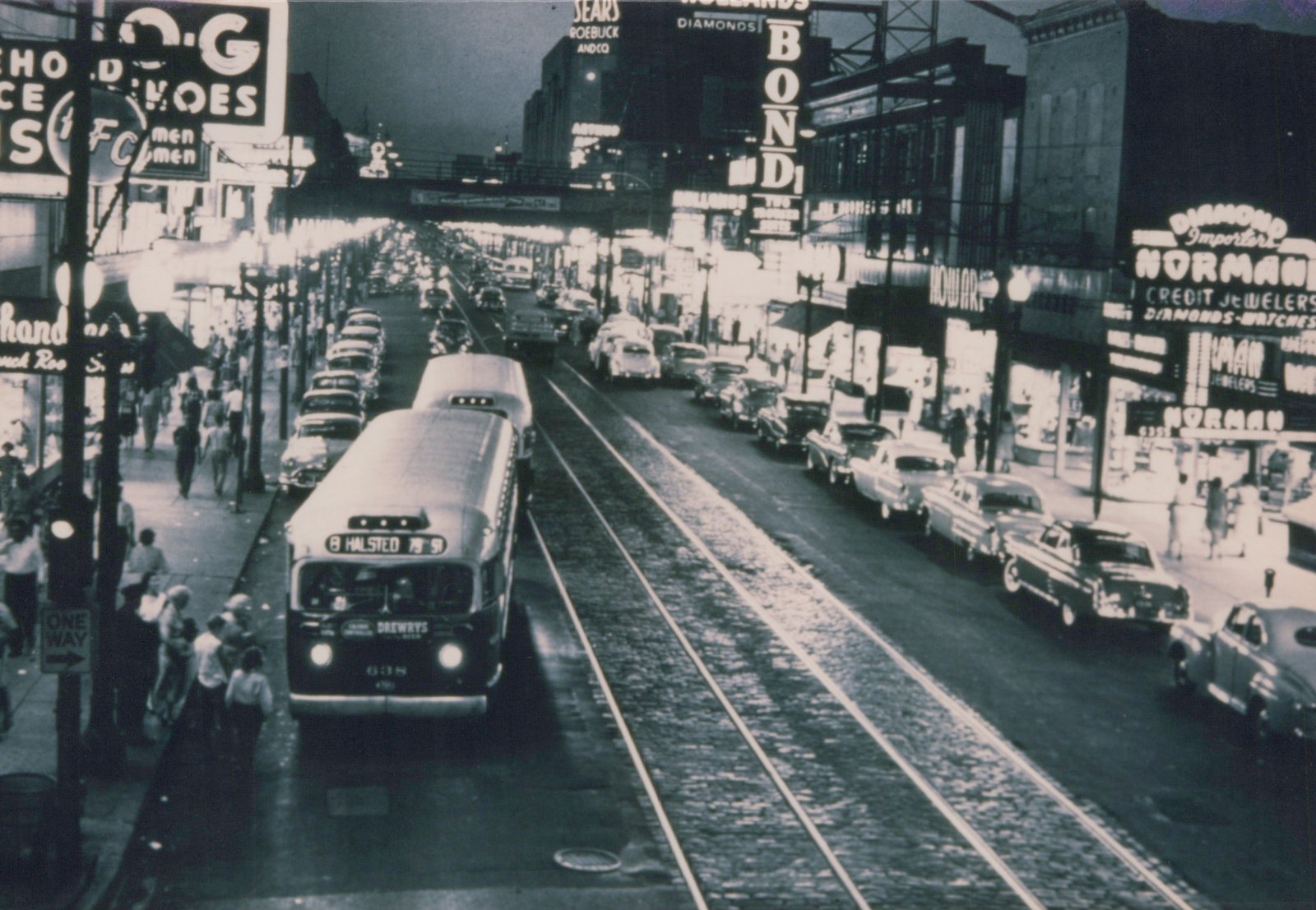 A black and white photo shows a CTA bus on a city street at dusk, surrounded by parked cars and pedestrians, with large glowing signs for shops like Sears and Bond's above.