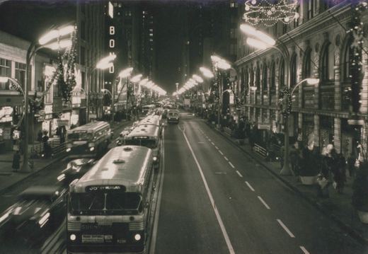 Nighttime street scene with a CTA bus on State Street in Chicago, surrounded by holiday lights, pedestrians, and other traffic.
