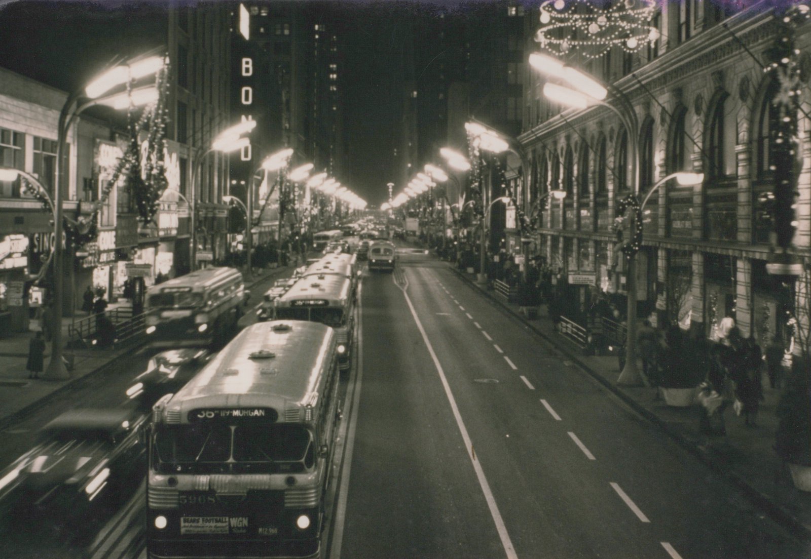 Nighttime street scene with a CTA bus on State Street in Chicago, surrounded by holiday lights, pedestrians, and other traffic.