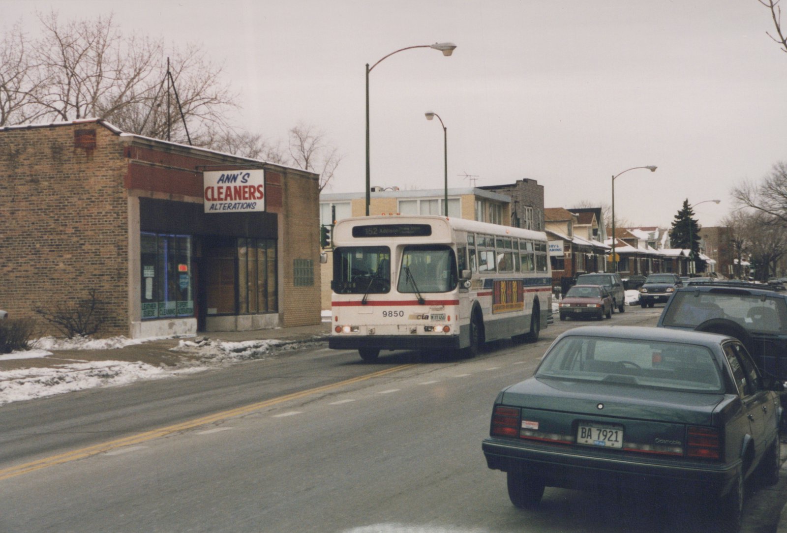 A white Chicago Transit Authority bus with the number 9850 is driving down a snowy street, with a green Oldsmobile and other cars nearby, and a store named Ann's Cleaners visible on the left.